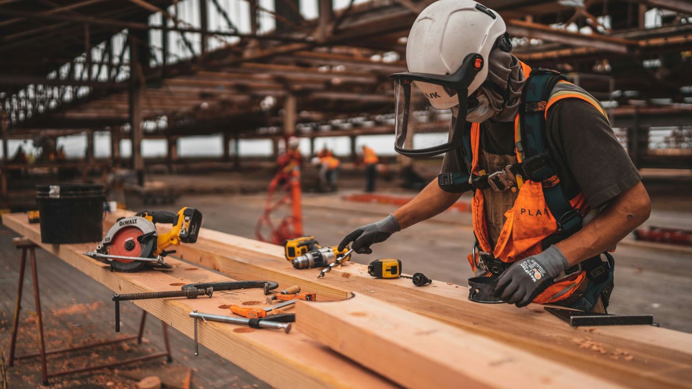A construction worker in a hard hat and face shield measuring lumber on a job site